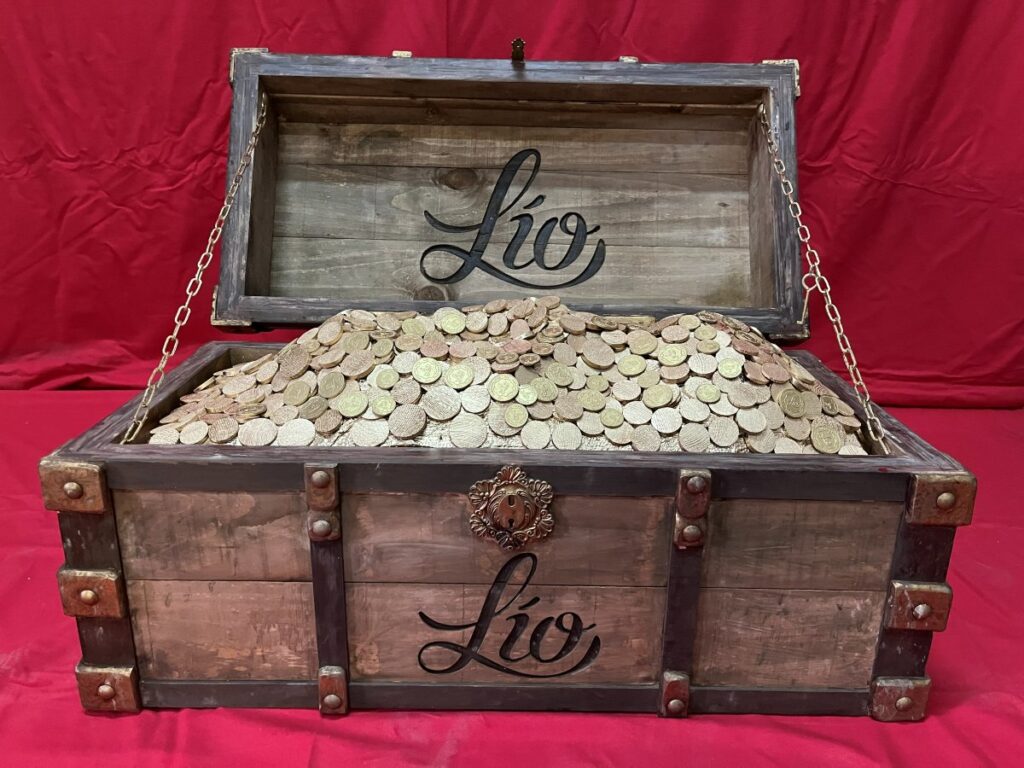 Cofre de madera envejecida con herrajes, abierto y lleno de monedas, con el logotipo de Lío
