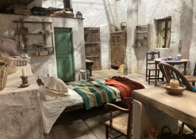 Interior de cabaña rural con paredes encaladas, puerta verde desconchada y cama con manta de rayas