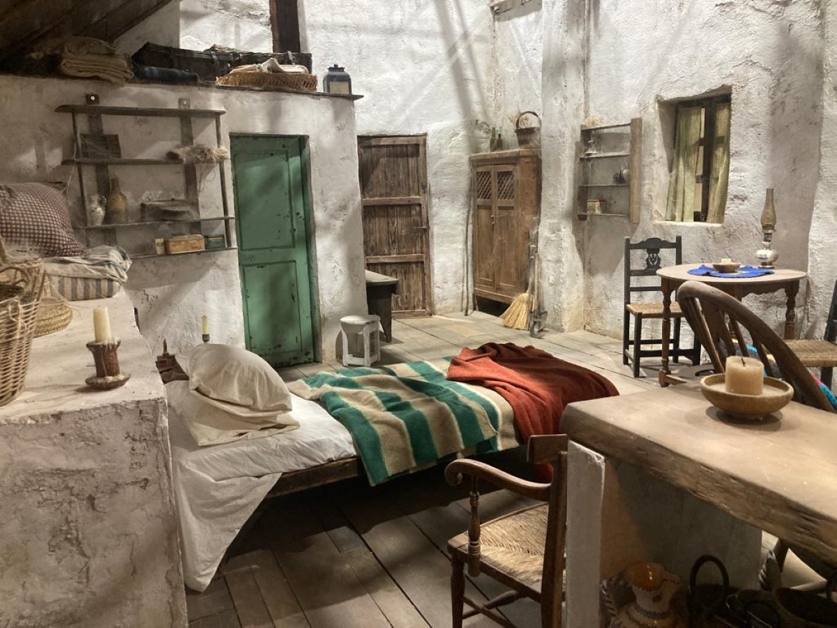 Interior de cabaña rural con paredes encaladas, puerta verde desconchada y cama con manta de rayas