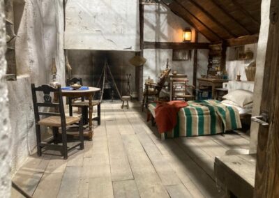 Interior de la cabaña con suelo de tablones, mesa con sillas de enea y cama