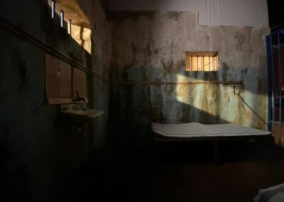 Interior de una celda con cama, lavabo y muro con manchas de humedad