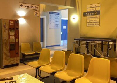 Sala de espera de hospital con sillas amarillas, máquina expendedora y carteles de planta