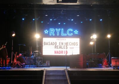 Escenario con el cartel luminoso «Basado en hechos reales» y el rótulo RYLC en neón azul
