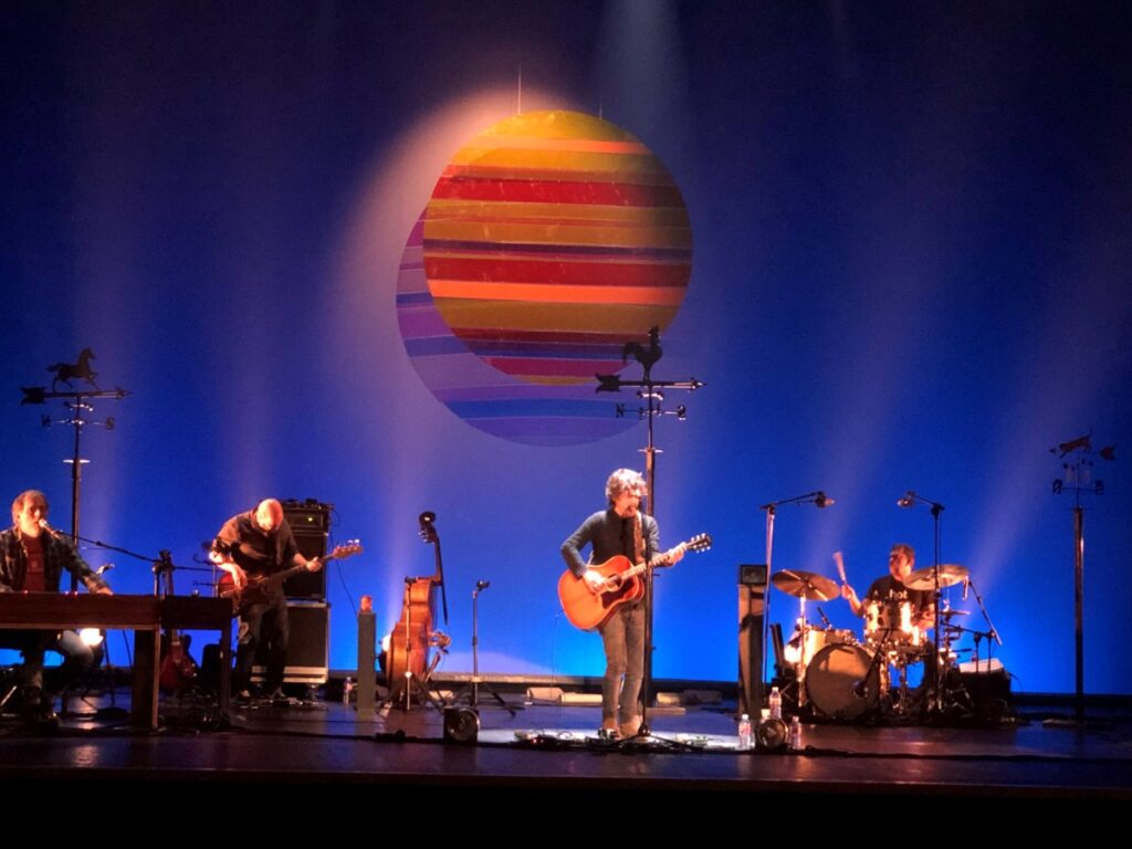 Quique González en concierto con un gran sol de franjas de colores suspendido sobre el escenario