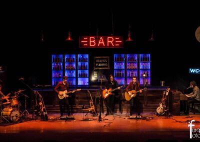 Escenario con barra de bar, botelleros iluminados en azul y un letrero de neón BAR
