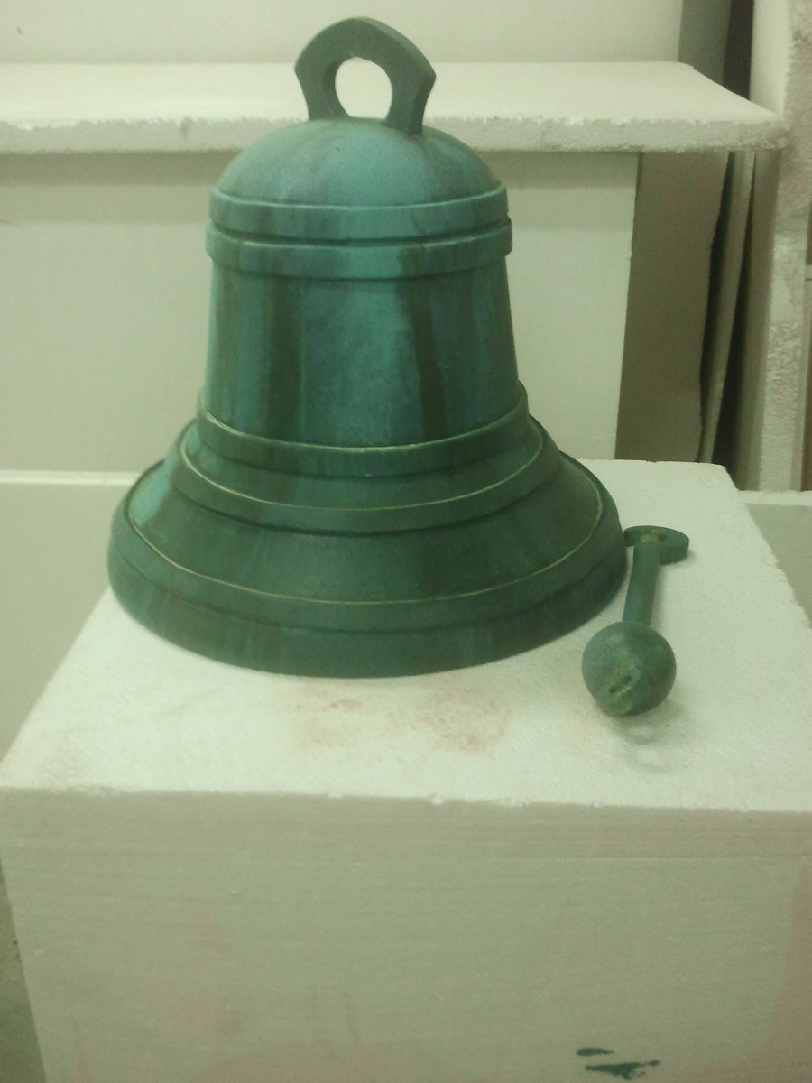 Campana de bronce verde ficticia con su badajo, sobre una peana blanca